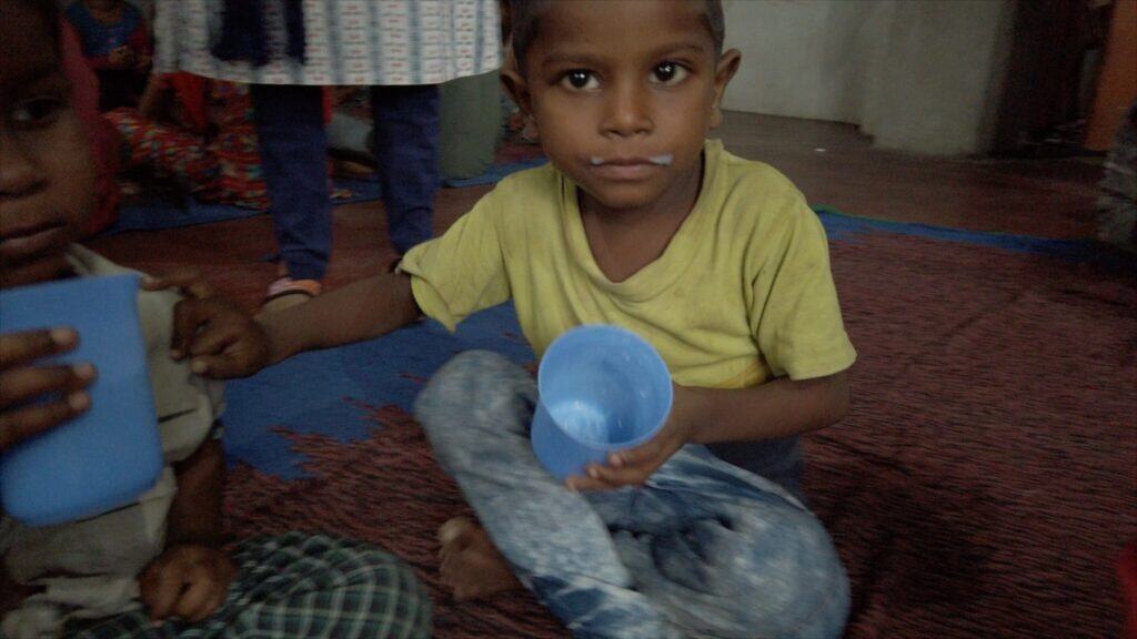 Zwei Kinder sitzen auf dem Boden und halten blaue Tassen mit Milch in den Händen. Ein Kind hat Milch auf der Oberlippe.