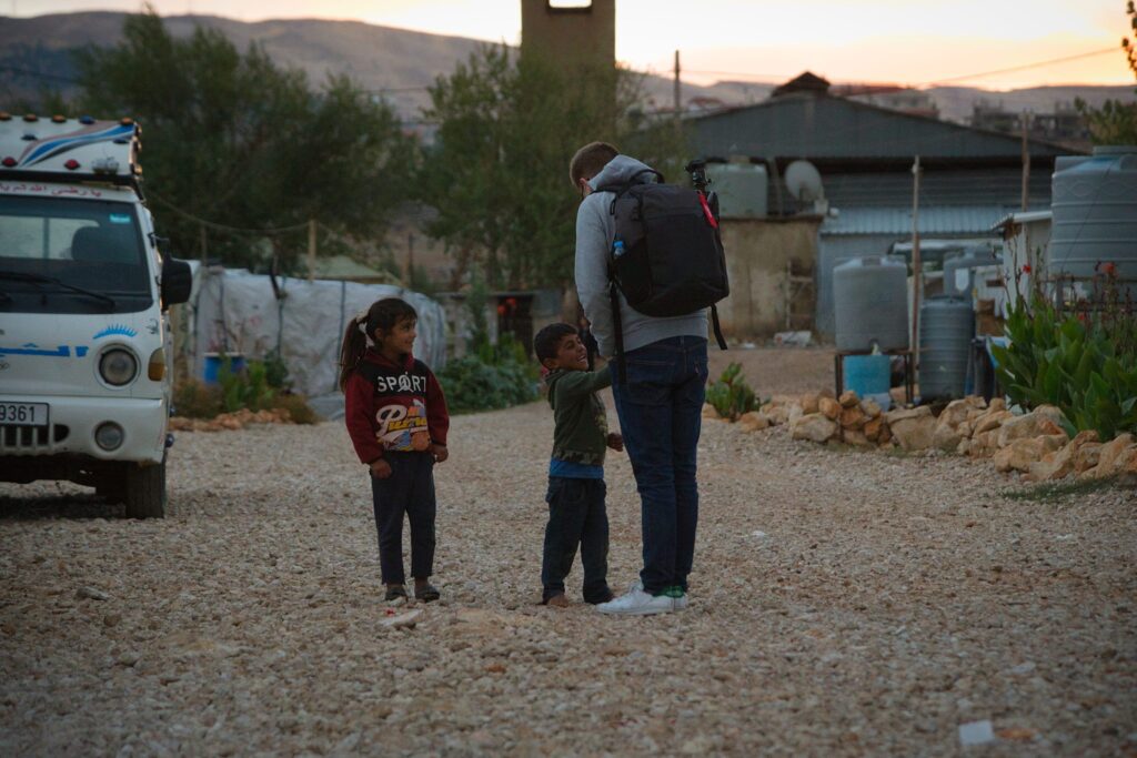 Mann mit Rucksack interagiert mit zwei Kindern in einem Flüchtlingslager.