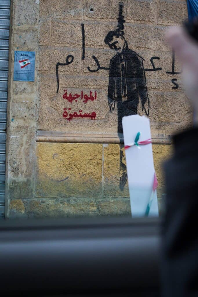 Graffiti, das eine hängende Figur an einer Steinmauer darstellt; arabischer Text sichtbar.