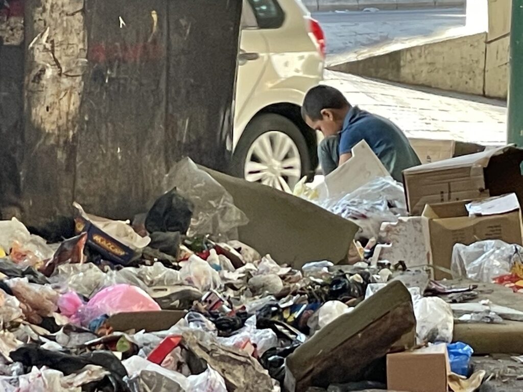 Kind sucht zwischen Müll und Schutt in der Nähe eines Müllcontainers nach Wertstoffen.