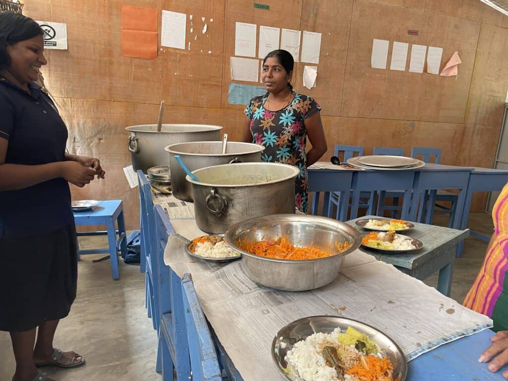 Zwei Frauen servieren Essen bei einem Schulmittagessenprogramm. Man sieht große Metalltöpfe und Teller mit Reis und Gemüse.
