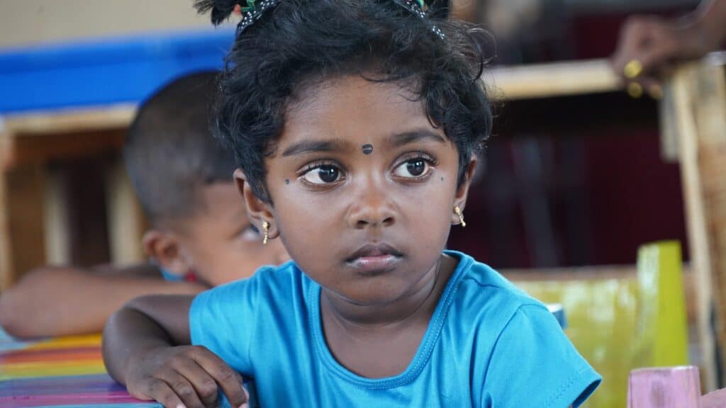 Ein junges Mädchen mit dunklem Haar und Bindi sowie einem blauen Hemd sitzt an einem bunten Tisch.