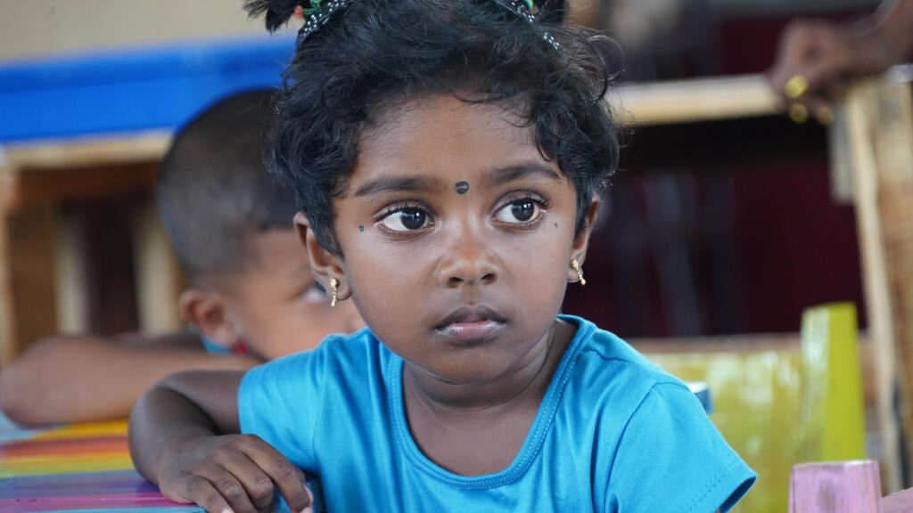 Ein junges Mädchen mit dunklem Haar und Bindi sowie einem blauen Hemd sitzt an einem bunten Tisch.