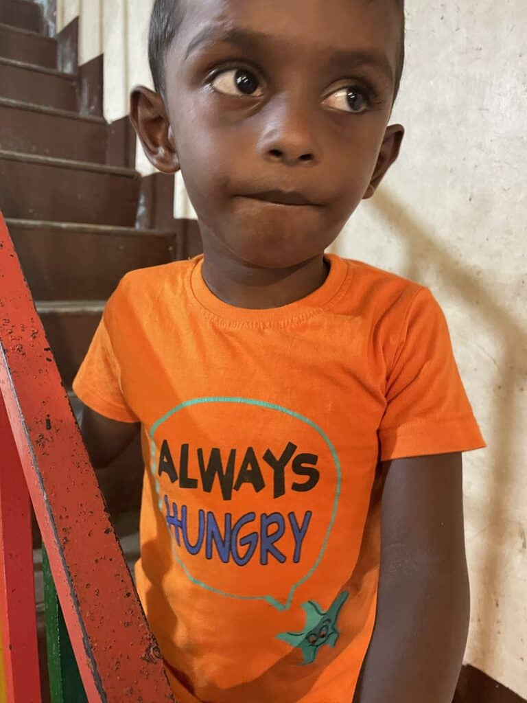 Kleiner Junge in einem orangefarbenen T-Shirt mit der Aufschrift „Always Hungry“ in der Nähe einer Treppe.