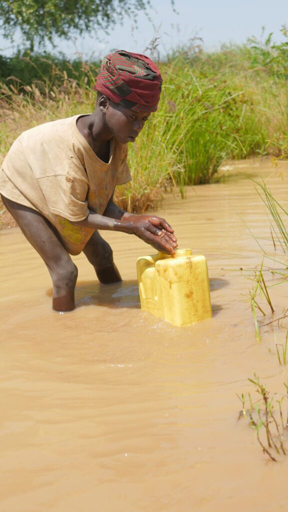 Kleines Kind füllt einen Wasserbehälter aus einem schlammigen Fluss.