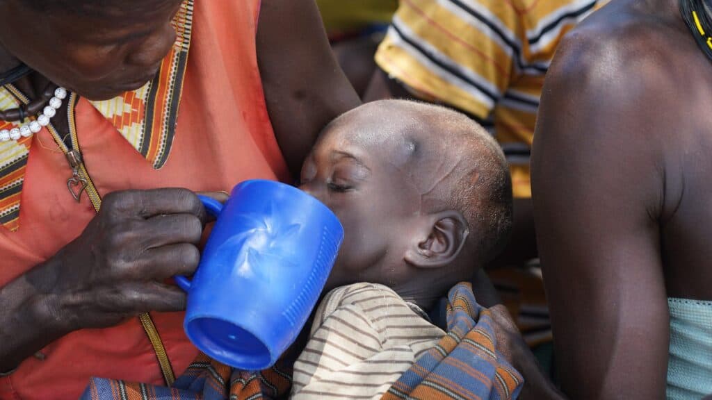 Eine ältere Frau füttert ein Kind mit Wasser aus einem blauen Becher.