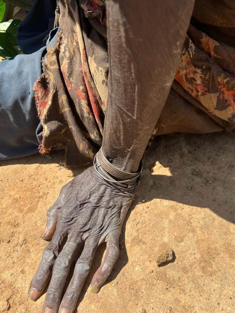 Nahaufnahme der mit Armbändern geschmückten Hand einer älteren Frau, die auf sonnengetrockneter Erde ruht.