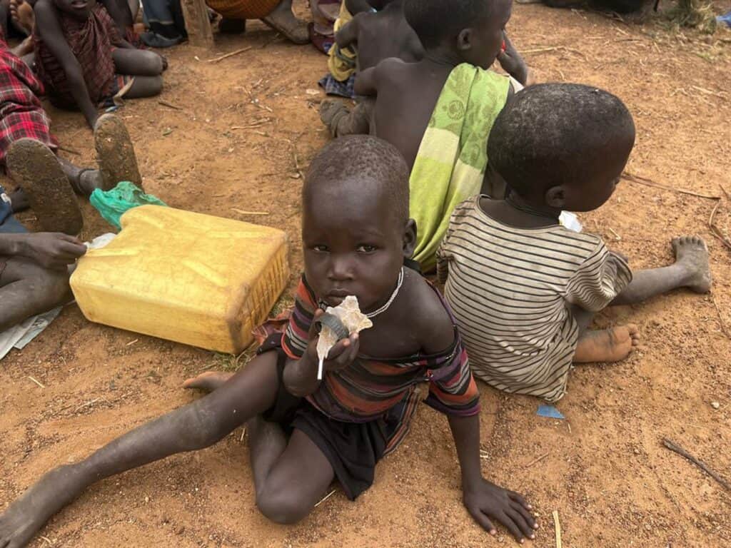 Unterernährte Kinder sitzen auf dem Boden, eines hält ein Stück Essen in der Hand.