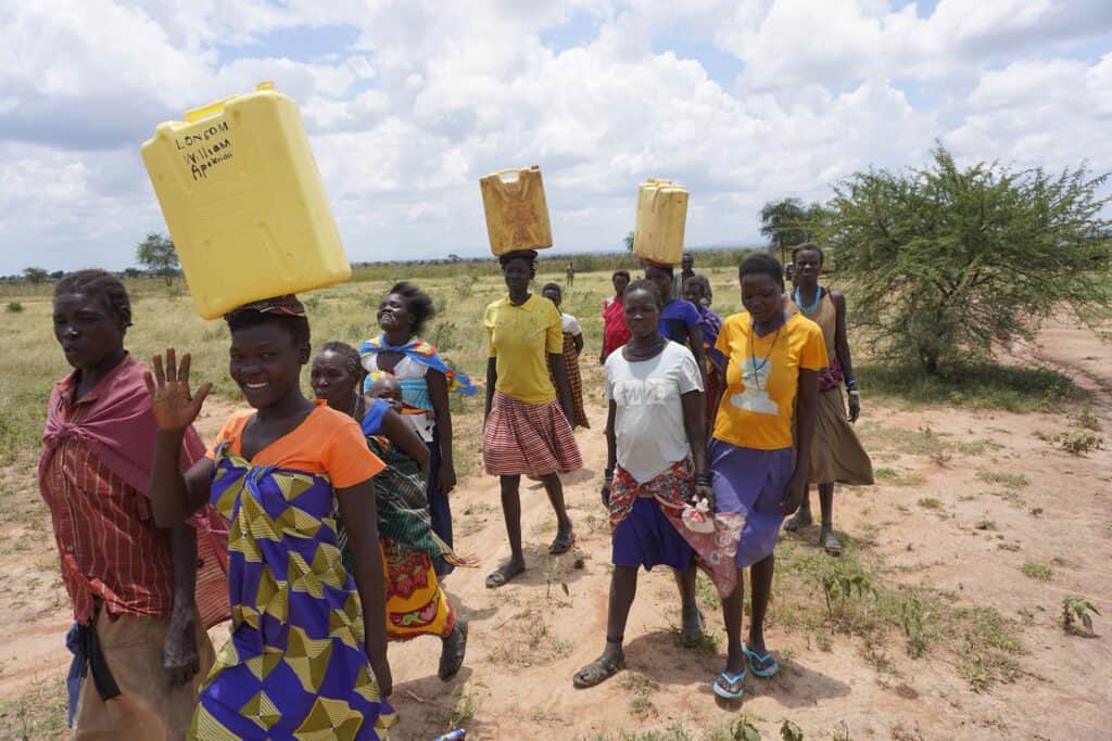Afrikanische Frauen tragen Wasserbehälter auf dem Kopf und gehen einen Feldweg entlang.