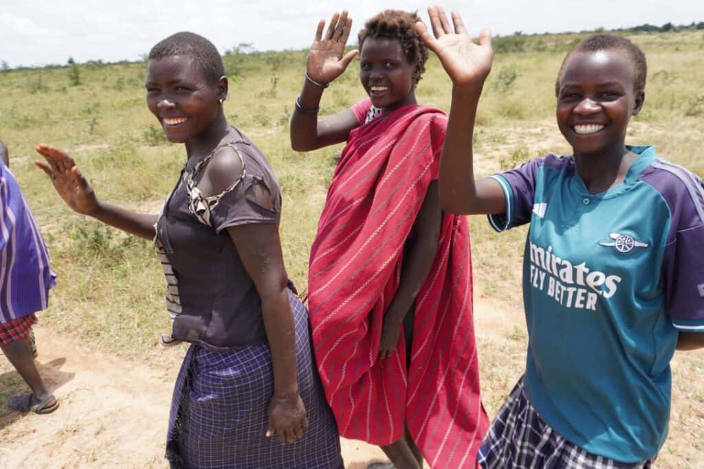 Drei lächelnde afrikanische Frauen in traditioneller und moderner Kleidung winken zur Begrüßung.