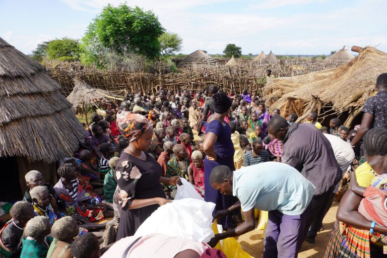 Helfer verteilen Hilfsgüter an eine große Gruppe von Menschen in einem ländlichen Dorf.