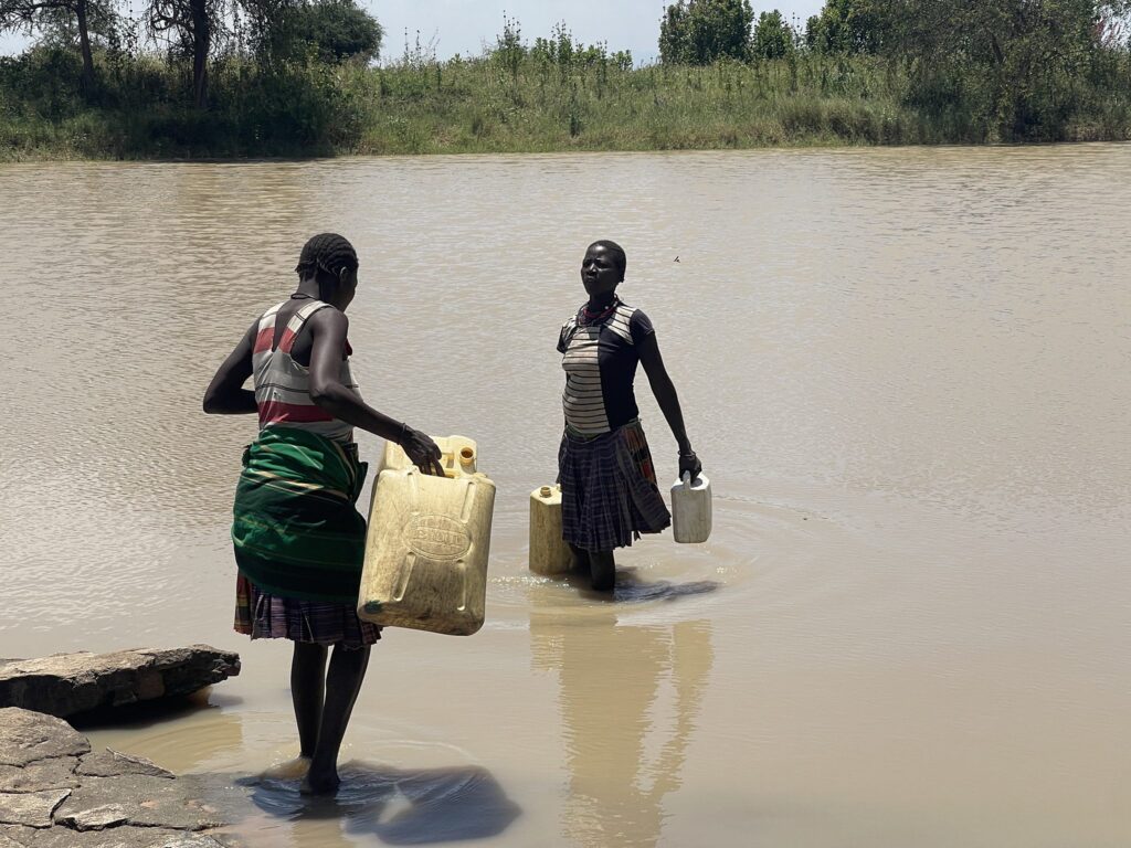 Zwei Frauen tragen Wasserbehälter und waten durch einen seichten Fluss.