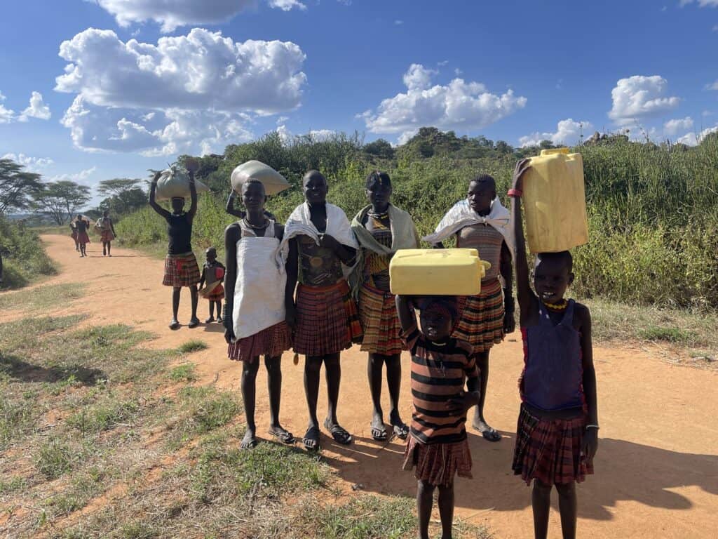 Eine Gruppe afrikanischer Frauen und Kinder trägt Wasser und Getreide auf einer unbefestigten Straße.