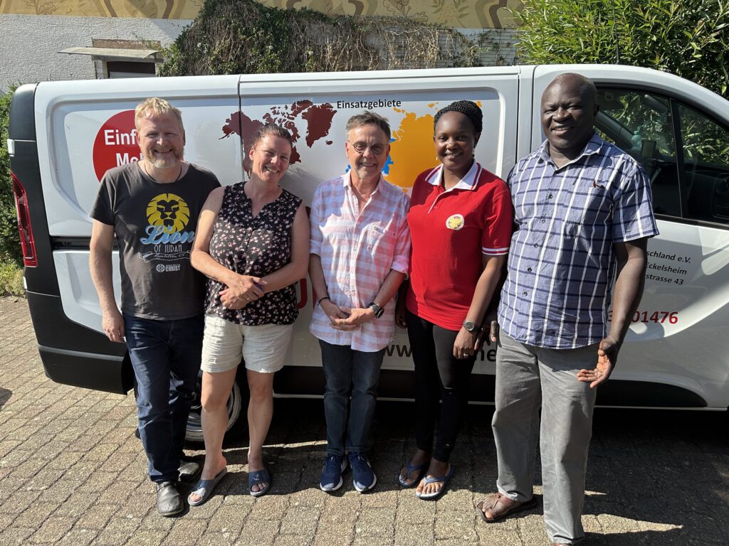 Ein Team aus fünf lächelnden Menschen steht vor einem weißen Lieferwagen, bereit für eine Missionsreise.