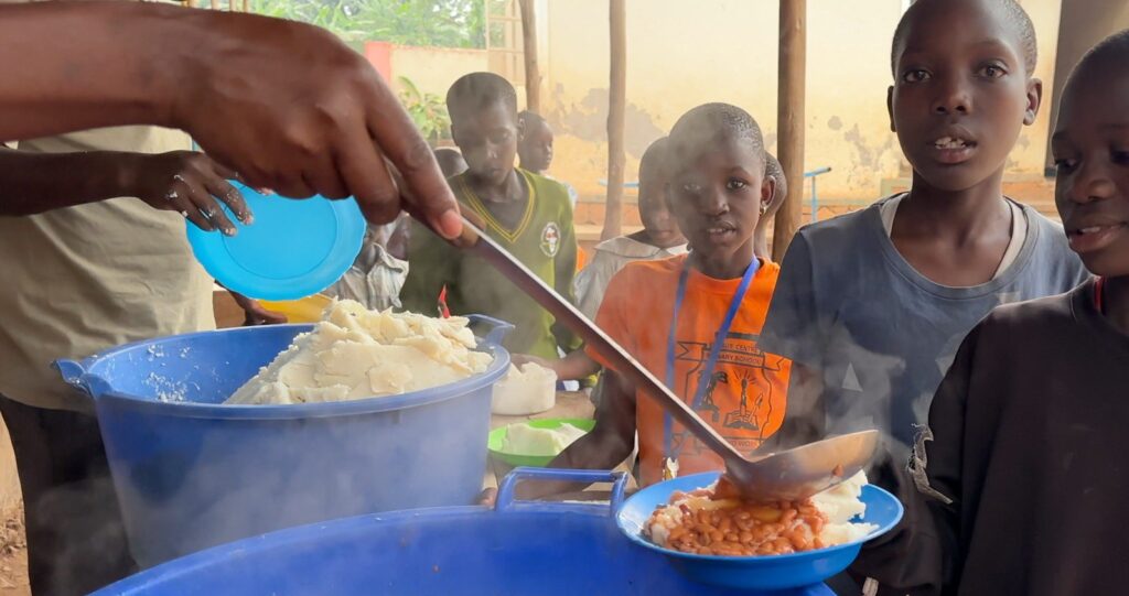 Freiwillige servieren Kindern im Rahmen eines Schulspeisungsprogramms eine warme Mahlzeit mit Bohnen und Posho.
