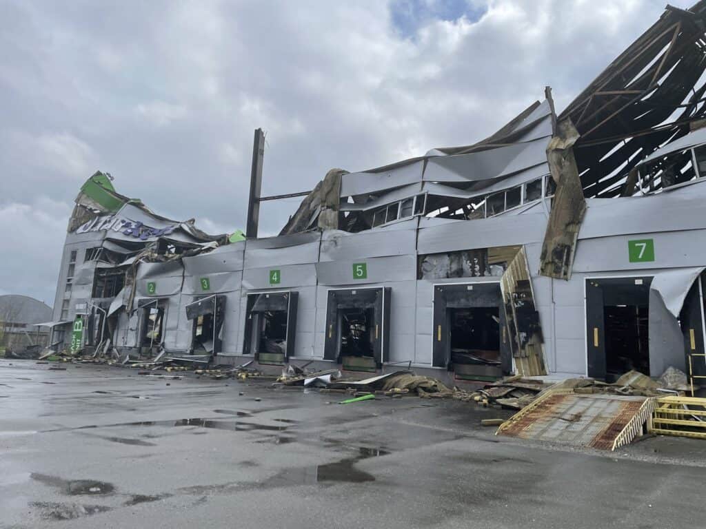 Beschädigtes Lagergebäude nach einem schweren Sturm mit erheblichen Dach- und Wandschäden. Die Nummern 2-7 sind auf den Laderampen sichtbar.