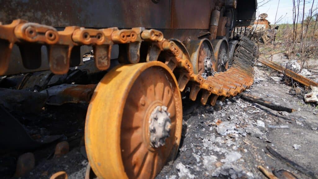 Nahaufnahme einer rostigen, ausgebrannten Panzerkette und eines Panzerrads. Kriegsschäden sind deutlich zu erkennen.