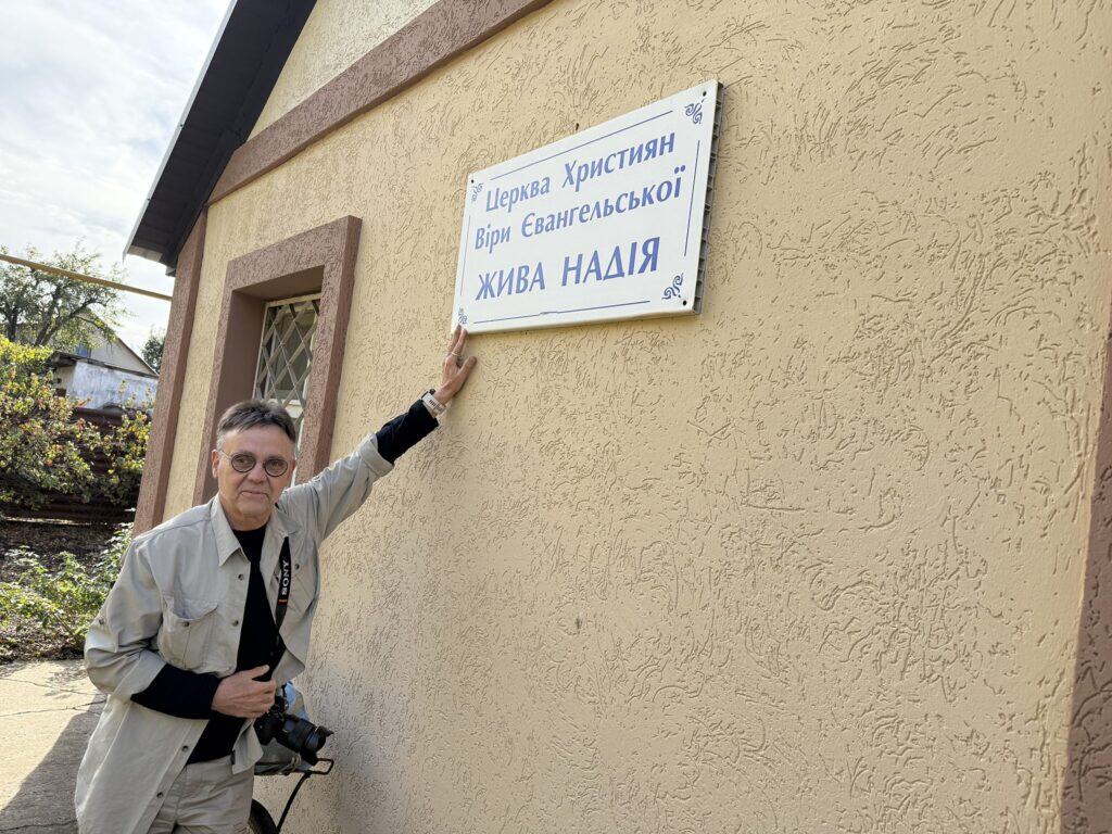 Ein Mann steht neben einem Kirchenschild mit der Aufschrift „Kirche der Christen evangelischen Glaubens, Lebendige Hoffnung“.