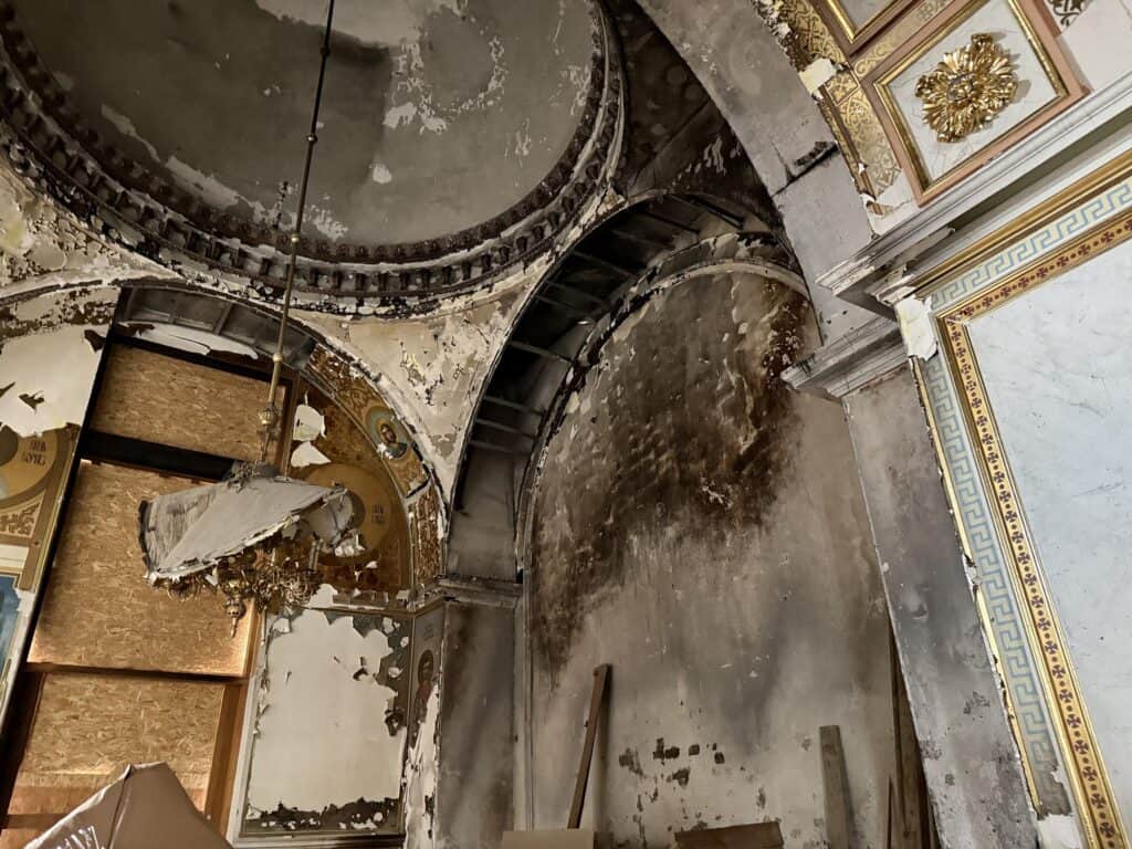 Beschädigtes Inneres einer historischen Kirche mit kunstvollem Stuck und verblassten Fresken. Ein Kronleuchter hängt unsicher.