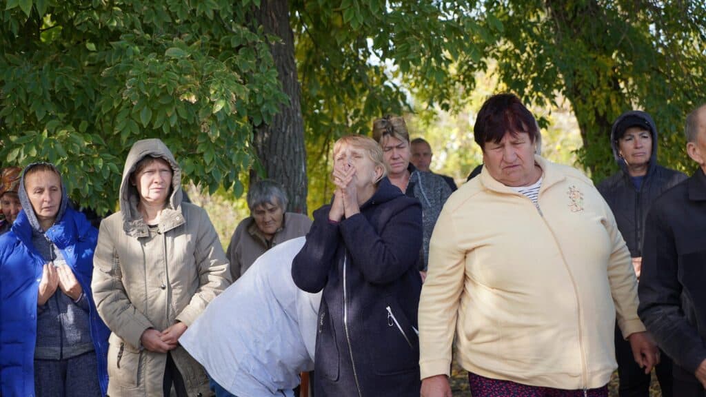 Trauernde versammeln sich mit gesenkten Köpfen an einer düsteren Gedenkstätte im Freien.