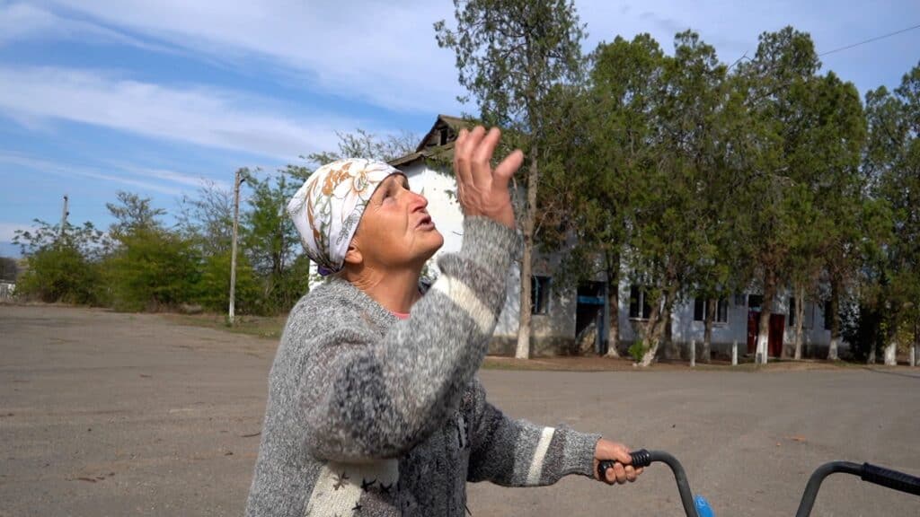 Ältere Frau blickt nach oben, möglicherweise betend, in der Nähe eines beschädigten Gebäudes