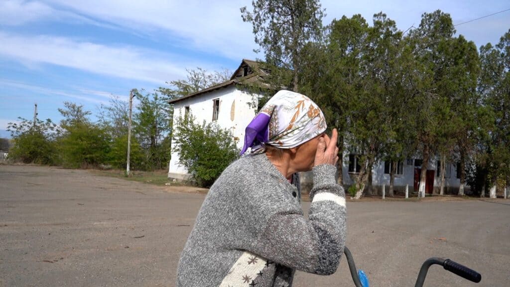 Ältere Frau mit Kopftuch in der Nähe eines beschädigten Gebäudes, wirkt verzweifelt.