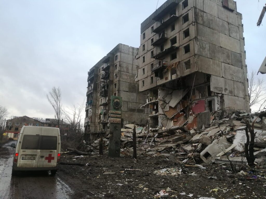 Zerstörte Wohnhäuser in der Ukraine; ein Krankenwagen parkt in der Nähe.