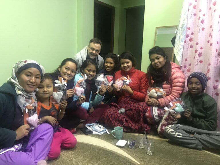 Eine Gruppe lächelnder Menschen, hauptsächlich Frauen und Kinder, hält handgemachte herzförmige Geschenke in den Händen. Hinter ihnen sitzt ein Mann.