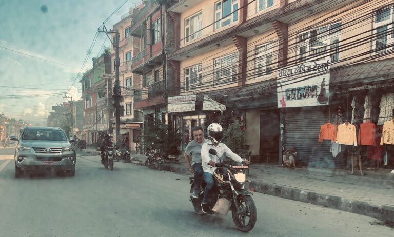 Motorradfahrer mit Beifahrer auf einer belebten nepalesischen Straße, vorbei an Geschäften und Autos.