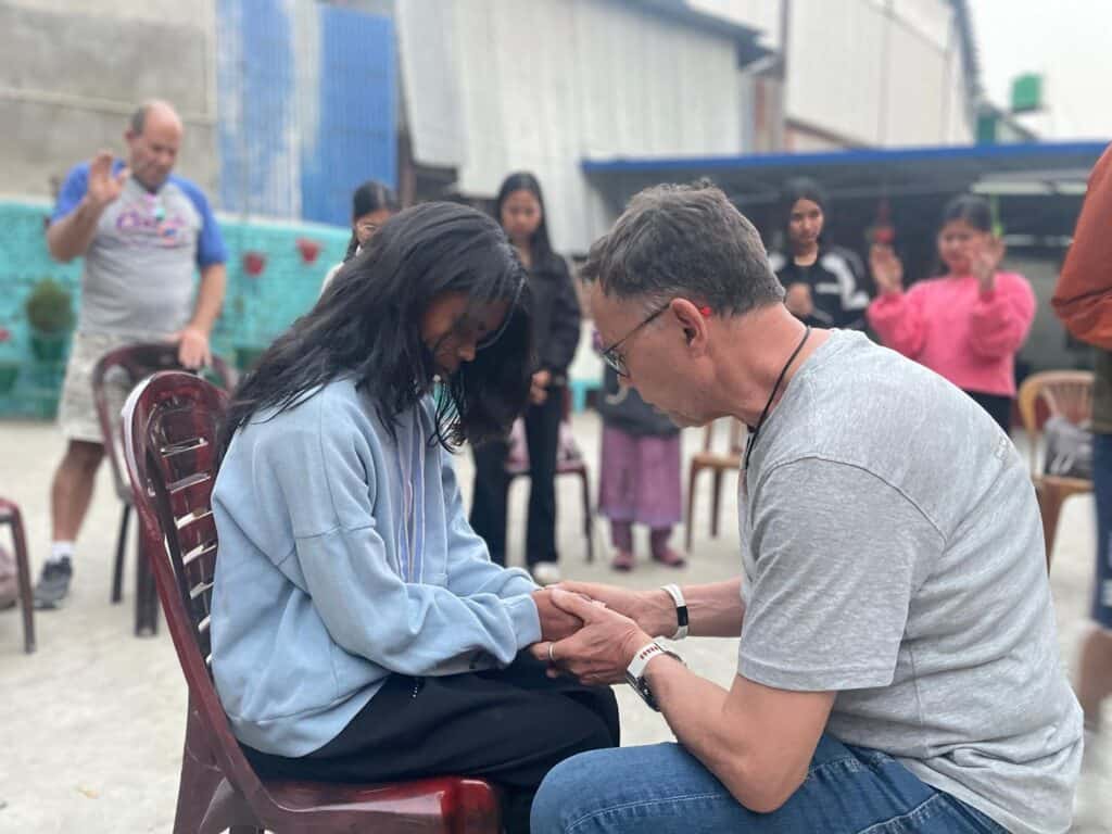 Mann betet mit junger Frau, die Hände gefaltet. Gruppe im Hintergrund.