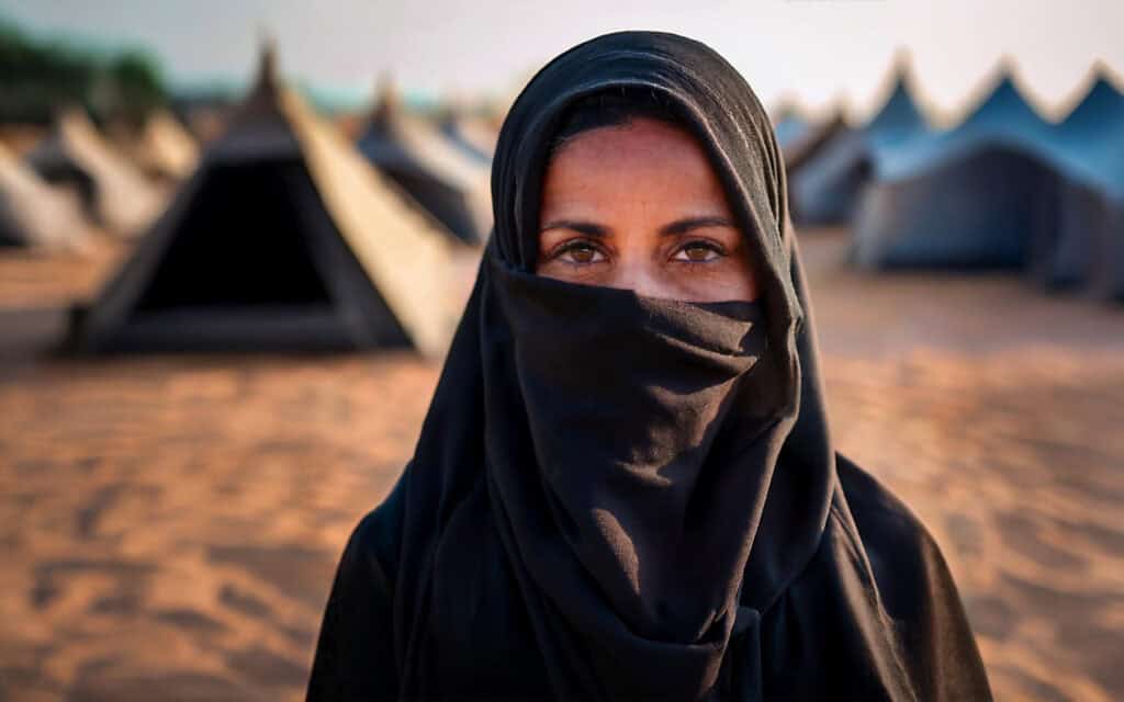 Eine Frau mit einem schwarzen Kopftuch steht vor einer Reihe von Zelten auf sandigem Boden. Ihr Gesicht ist größtenteils bedeckt, nur ihre Augen sind zu sehen. Der Hintergrund ist unscharf und zeigt weitere Zelte im Tageslicht.