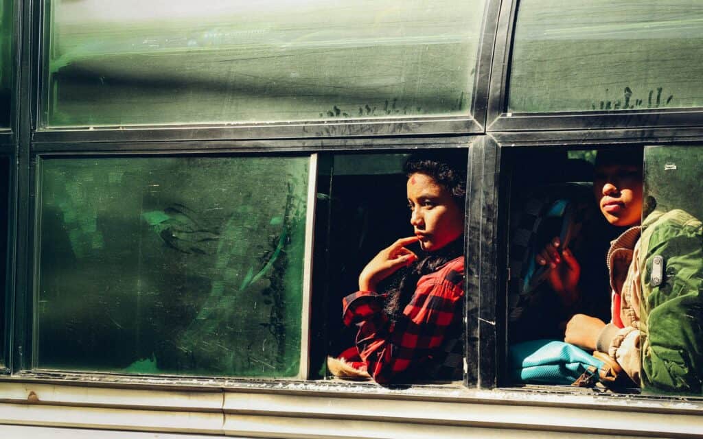 Zwei Personen sitzen in einem Bus und schauen aus einem offenen Fenster. Das Sonnenlicht beleuchtet teilweise ihre Gesichter, und eine Person trägt ein rotkariertes Hemd und stützt ihr Kinn auf die Hand. Das Busfenster ist staubig.