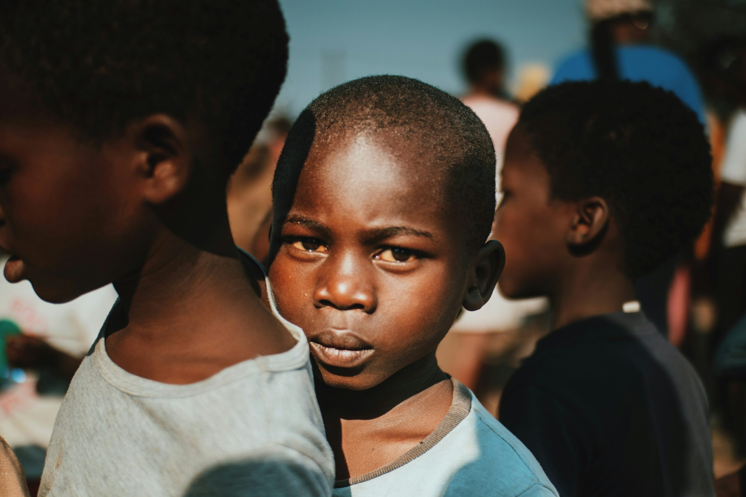 Ein kleiner Junge schaut direkt in die Kamera, während er zwischen anderen Kindern im Freien in hellem Sonnenlicht steht, mit Schatten und verschwommenen Figuren im Hintergrund.