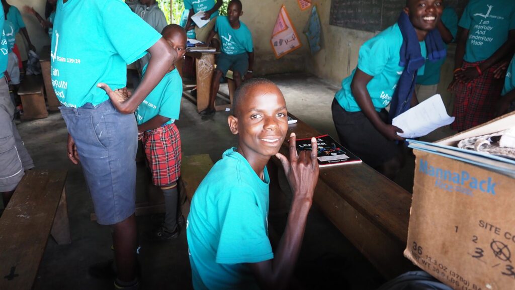 Lächelnder afrikanischer Junge im blaugrünen T-Shirt macht Peace-Zeichen im Klassenzimmer