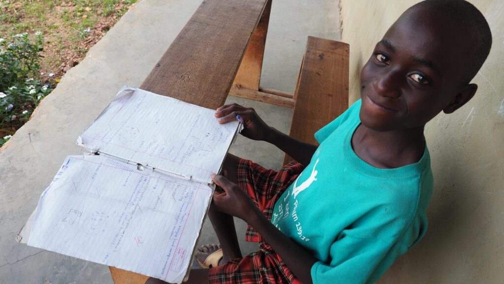 Lächelnder afrikanischer Junge studiert Matheaufgaben in abgenutztem Notizbuch an rustikalem Schreibtisch.