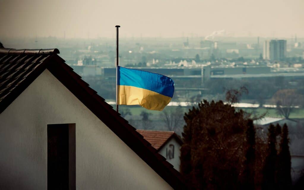 Eine ukrainische Flagge weht auf Halbmast auf einem Wohnhausdach, mit einer Stadtlandschaft und einem dunstigen Himmel im Hintergrund, was eine düstere oder trauernde Atmosphäre vermittelt.