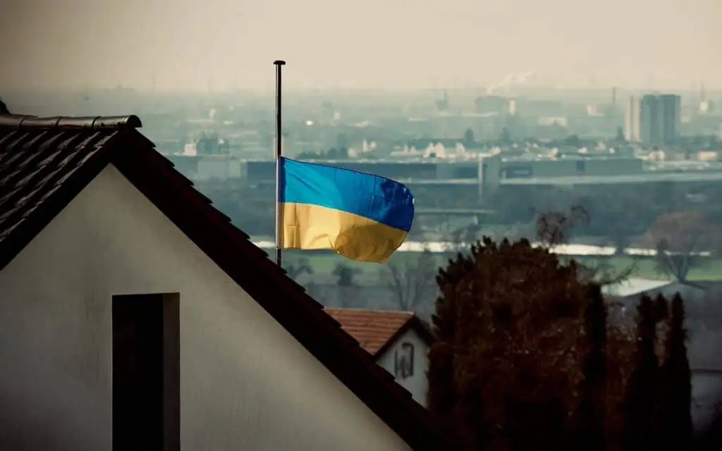 Eine ukrainische Flagge weht auf Halbmast auf einem Wohnhausdach, mit einer Stadtlandschaft und einem dunstigen Himmel im Hintergrund, was eine düstere oder trauernde Atmosphäre vermittelt.