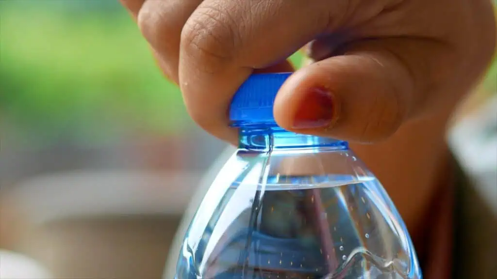 Nahaufnahme einer Hand mit rotem Nagellack, die den blauen Deckel einer mit Wasser gefüllten klaren Plastikflasche abschraubt. Der Hintergrund ist unscharf mit grünen und neutralen Tönen.