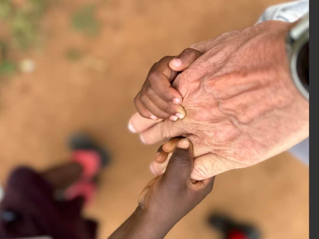 Eine Nahaufnahme von zwei kleinen dunkelhäutigen Händen, die sanft die faltige Hand einer älteren hellhäutigen Person halten, was Fürsorge und Verbindung symbolisiert. Der Hintergrund besteht aus verschwommener Erde und einer Person in rosa Schuhen.