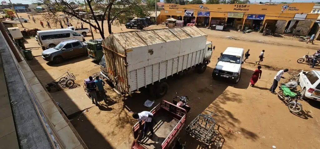 Eine Straßenszene zeigt einen großen weißen Lastwagen, der neben mehreren Fahrzeugen und Motorrädern vor gelben Schaufenstern geparkt ist. An einem sonnigen Tag versammeln sich Menschen um den Lastwagen und die Geschäfte.