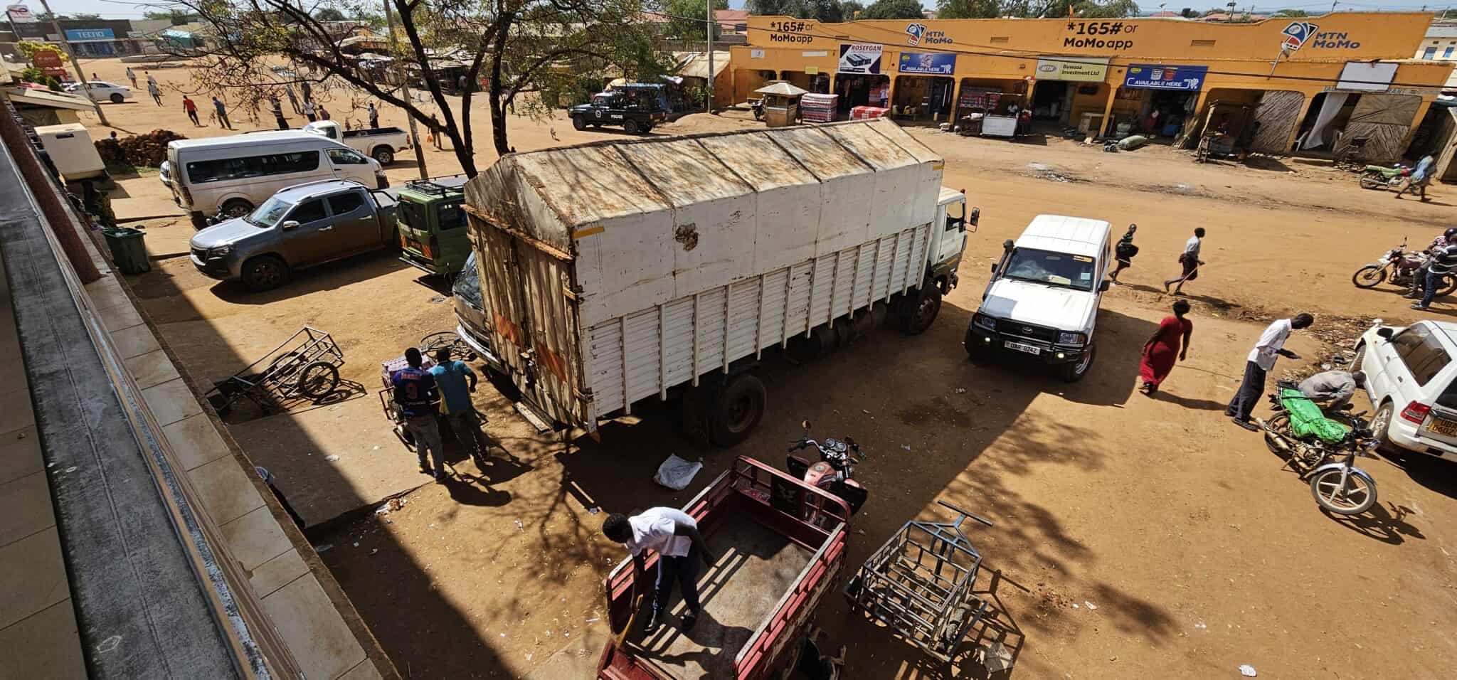 Eine Straßenszene zeigt einen großen weißen Lastwagen, der neben mehreren Fahrzeugen und Motorrädern vor gelben Schaufenstern geparkt ist. An einem sonnigen Tag versammeln sich Menschen um den Lastwagen und die Geschäfte.