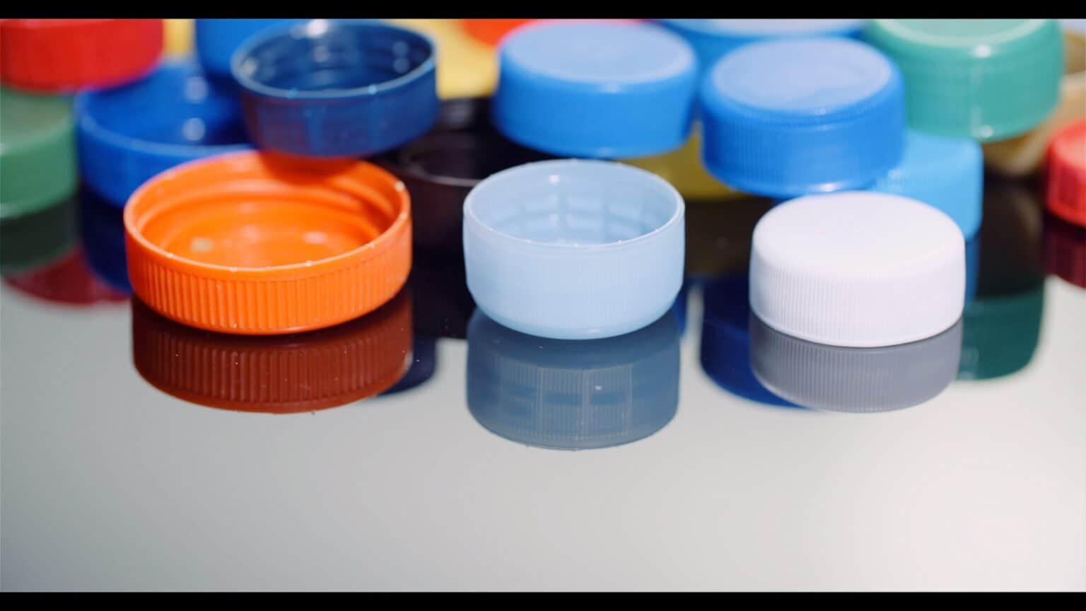 Eine Sammlung von bunten Plastikflaschenverschlüssen, darunter blau, orange, hellblau, weiß, grün und rot, die auf einer reflektierenden Oberfläche angeordnet sind.