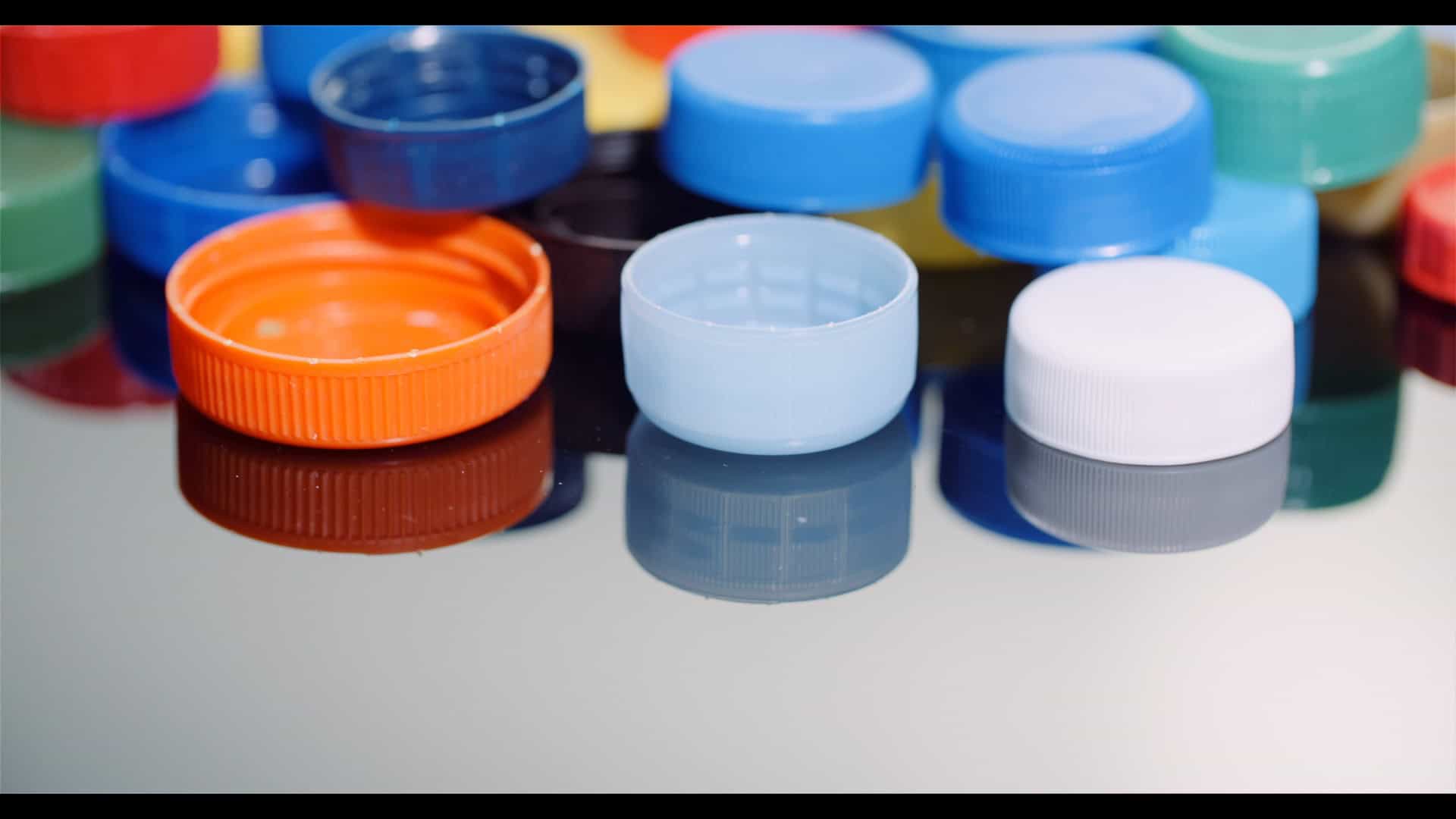 Eine Sammlung von bunten Plastikflaschenverschlüssen, darunter blau, orange, hellblau, weiß, grün und rot, die auf einer reflektierenden Oberfläche angeordnet sind.