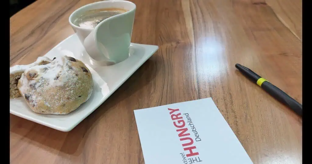 Auf einem Holztisch stehen eine Tasse Kaffee und ein Puderzuckergebäck auf einem weißen Teller. In der Nähe sind ein Stift und eine Karte mit der Aufschrift "THE HUNGRY Deutschland" zu sehen.