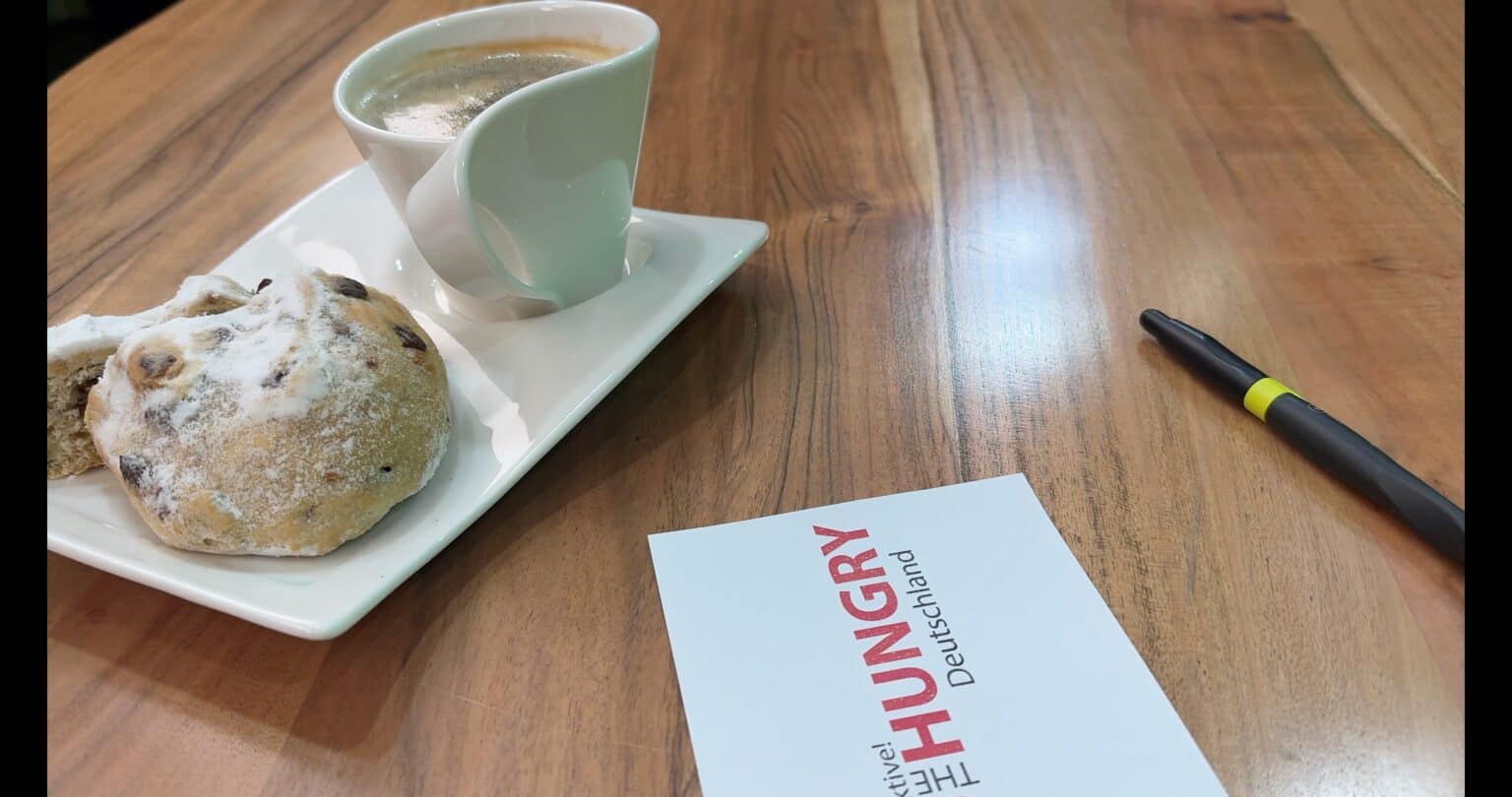 Auf einem Holztisch stehen eine Tasse Kaffee und ein Puderzuckergebäck auf einem weißen Teller. In der Nähe sind ein Stift und eine Karte mit der Aufschrift "THE HUNGRY Deutschland" zu sehen.