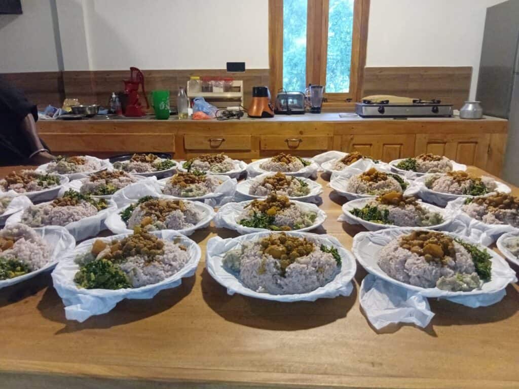 Teller mit Reis, Gemüse und einem Eintopf sind in Reihen auf einer hölzernen Arbeitsplatte in einer Küche angeordnet, bereit zum Servieren. Küchengeräte und Utensilien sind im Hintergrund zu sehen.