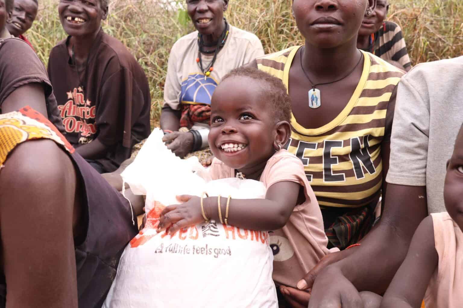 Ein lächelndes Kind umarmt einen großen weißen Sack mit Lebensmitteln, während es inmitten einer Gruppe von Erwachsenen und Kindern im Freien sitzt. Die Gruppe wirkt glücklich und entspannt, mit hohem Gras im Hintergrund.