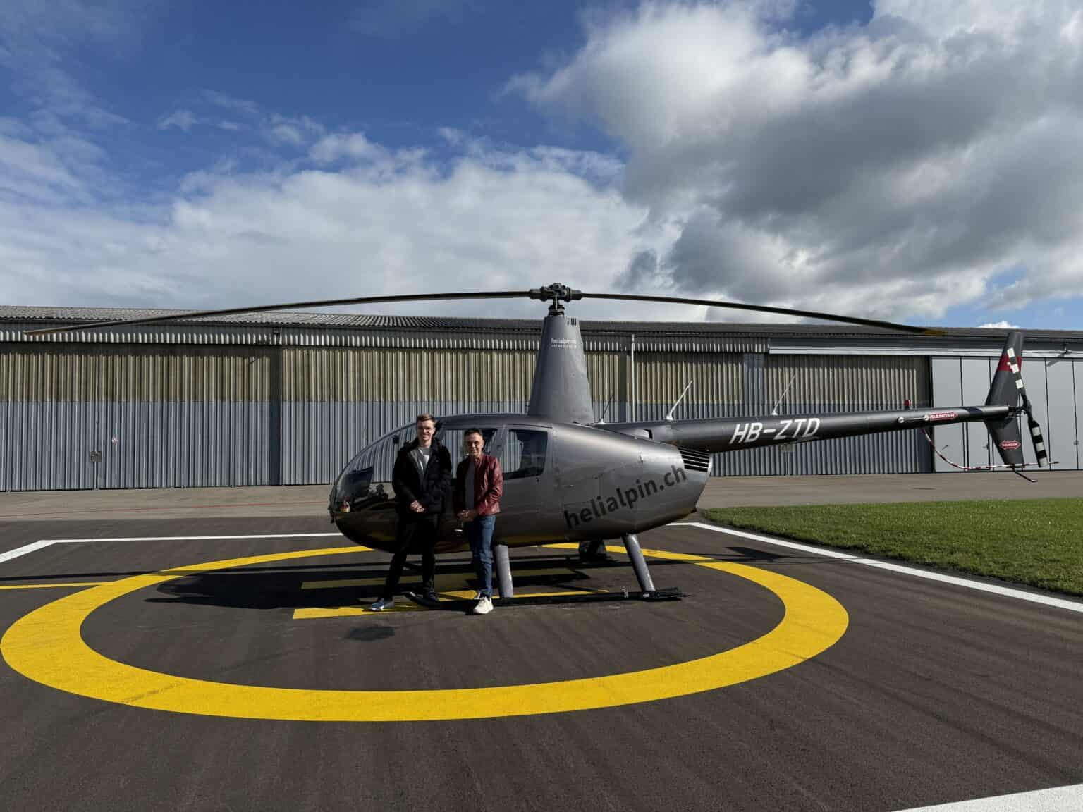 Zwei Personen stehen auf einem Hubschrauberlandeplatz neben einem grauen Helikopter mit der Aufschrift "helialpin.ch" vor einem Hangar unter einem teilweise bewölkten Himmel.