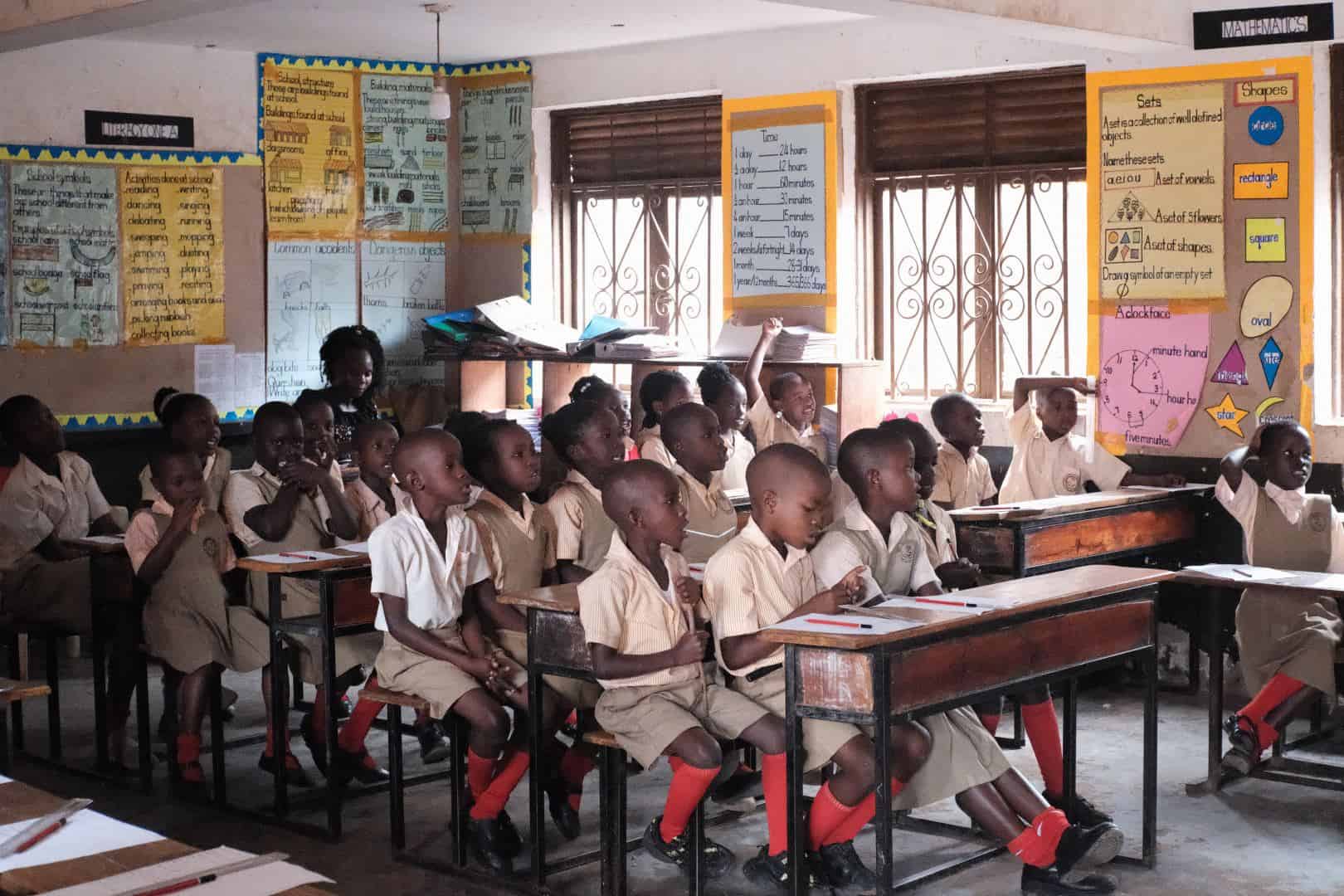 Ein Klassenzimmer voller junger Schüler in Uniform, die an den Tischen sitzen und nach vorne schauen. An den Wänden hängen bunte Lernplakate, und durch die vergitterten Fenster strömt Sonnenlicht herein.
