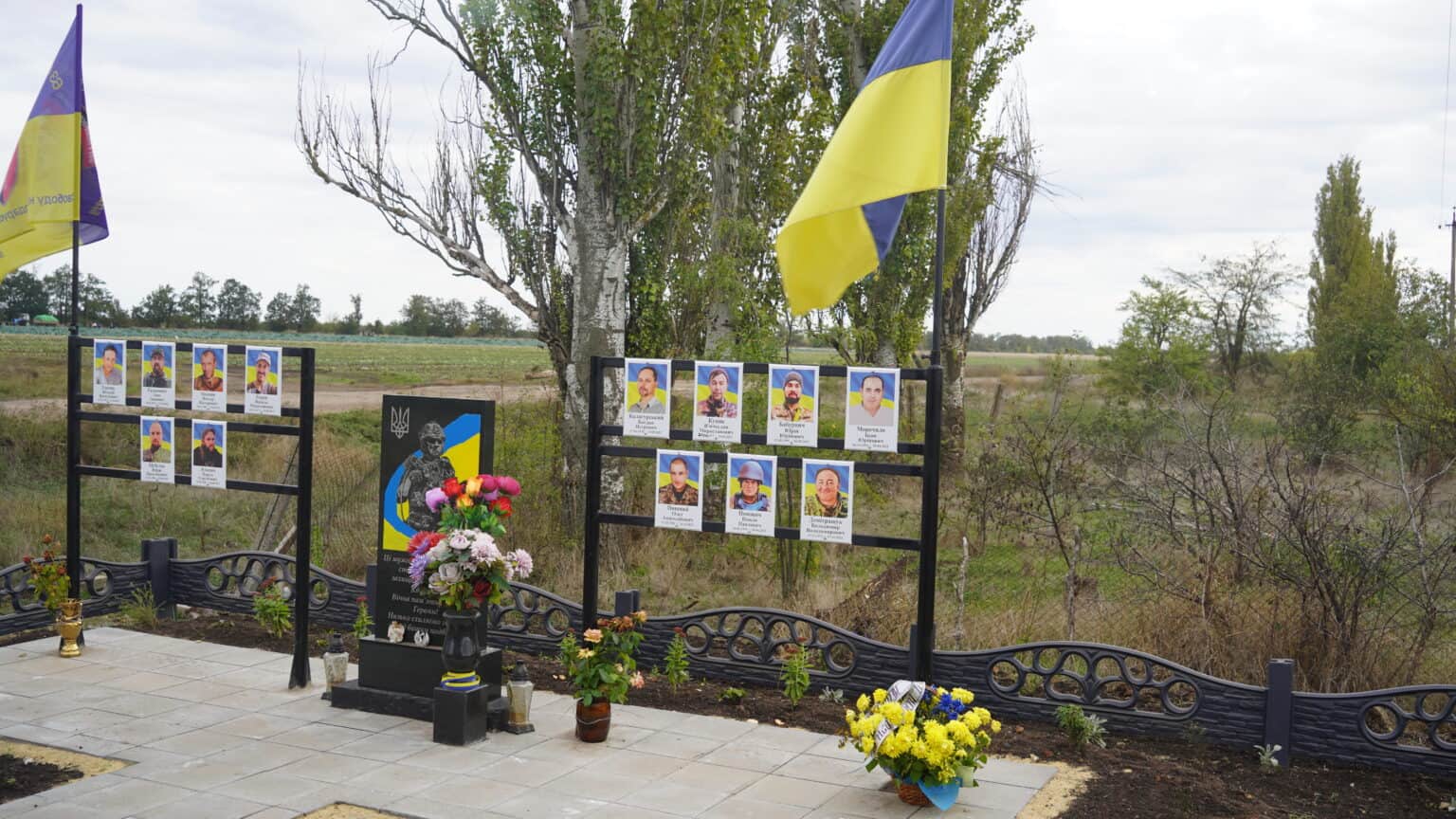 Eine Gedenkstätte im Freien mit ukrainischen Flaggen, Fotos von Einzelpersonen, Blumen und Gedenktafeln in einer ländlichen Landschaft mit Bäumen und einem bewölkten Himmel.